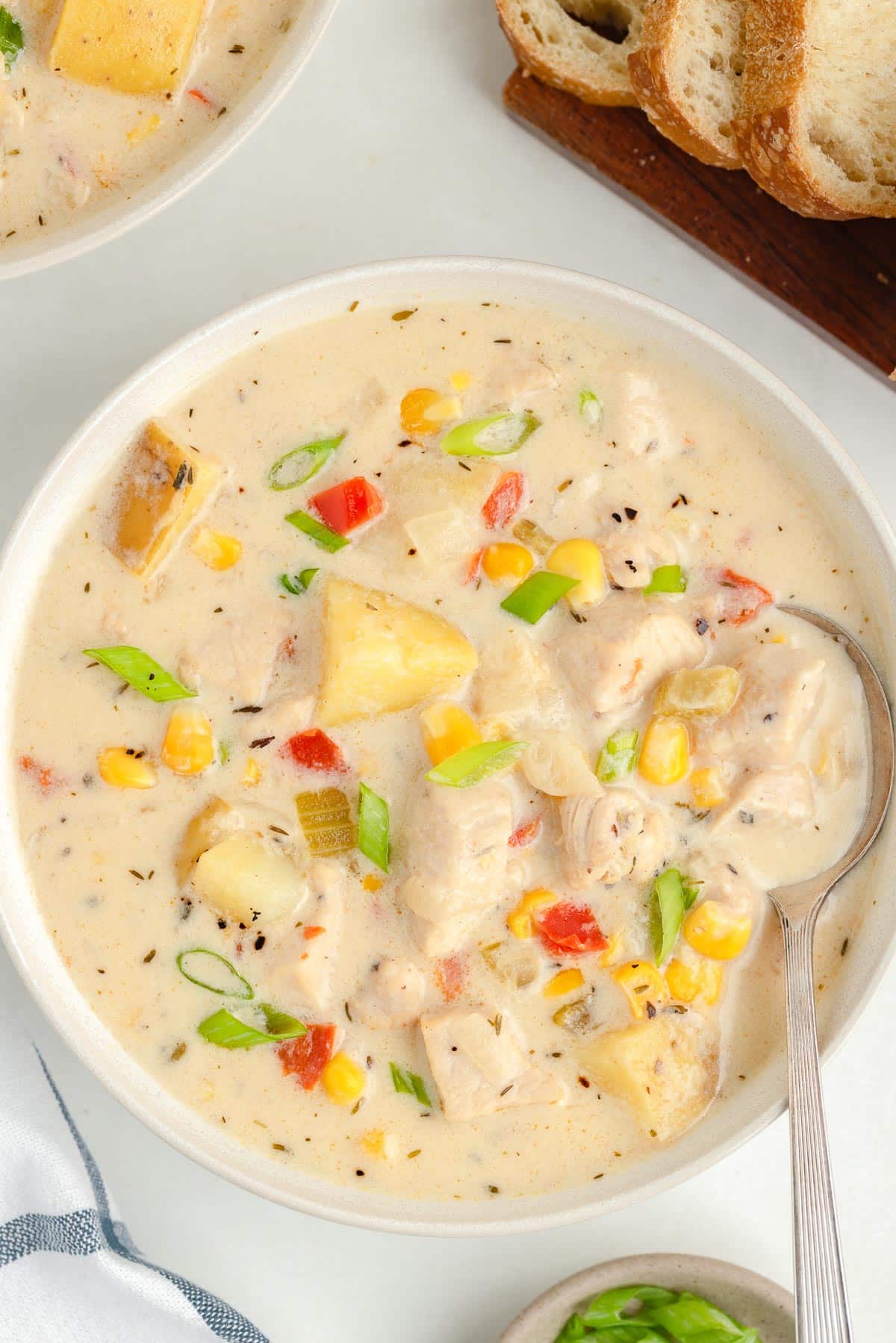 A bowl of creamy chicken and corn chowder with chunks of potatoes, corn, chicken and vegetables from overhead on a table with a spoon scooping and bread and a napkin nearby.