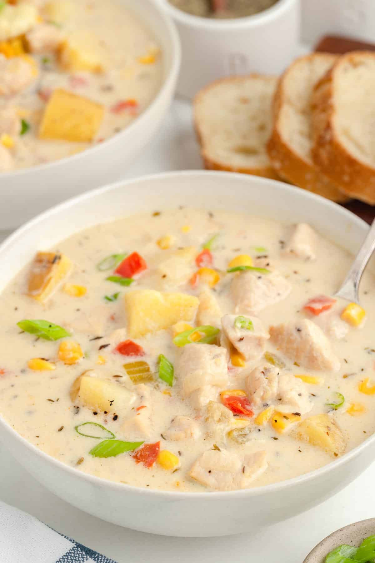 A bowl of creamy chicken corn chowder being stirred by a spoon with a second bowl of soup and sliced bread in the background.