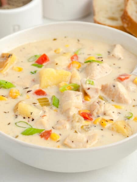 A bowl of creamy chicken corn chowder soup with chunks of potato, chicken, corn, green onion and bell pepper with a spoon and bread on a table.