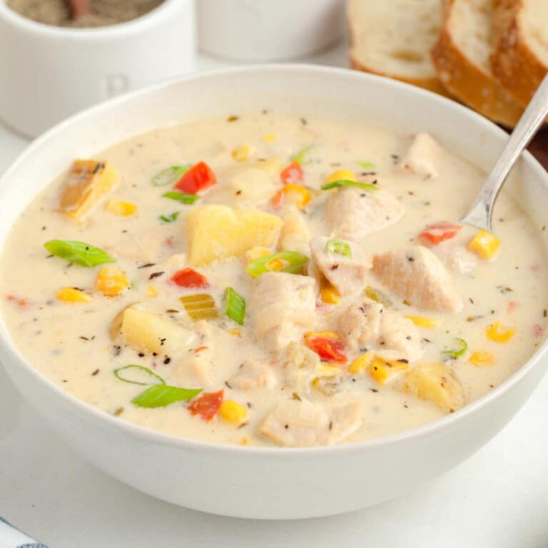 A bowl of creamy chicken corn chowder soup with chunks of potato, chicken, corn, green onion and bell pepper with a spoon and bread on a table.