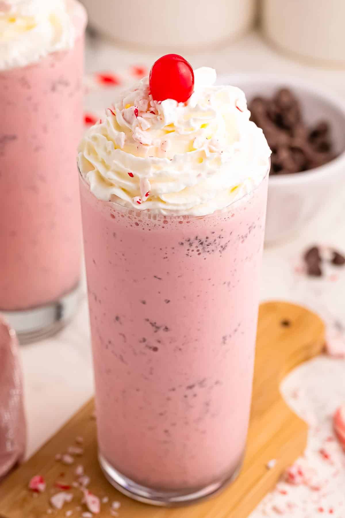 A talk glass full of pink chocolate chip peppermint milkshake, a copycat chick fil a recipe, topped with whipped cream, a maraschino cherry and crushed peppermint.