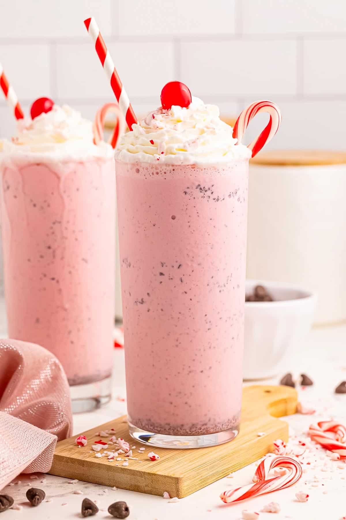 Two tall glasses of pink chocolate chip peppermint milkshake topped with whipped cream, a cherry from the side with red and white straws and a candy cane in the glass.