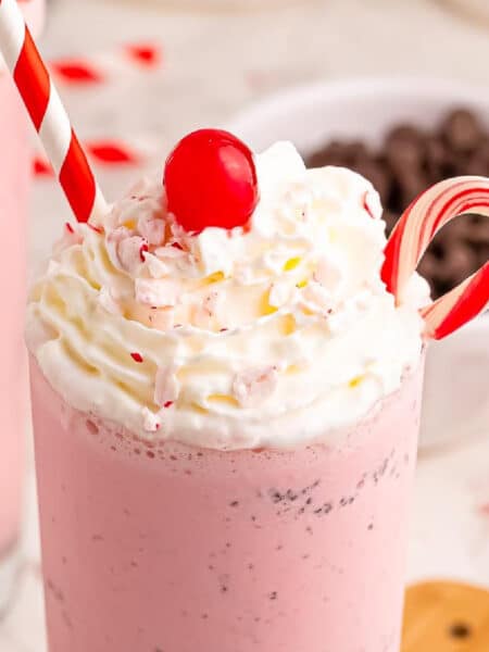 A pink milkshake topped with whipped cream, a red cherry, a candy cane, and a red-striped straw, with a bowl of chocolate chips in the background.