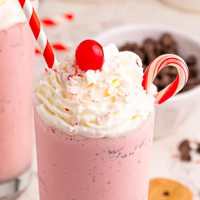 A pink milkshake topped with whipped cream, a red cherry, a candy cane, and a red-striped straw, with a bowl of chocolate chips in the background.