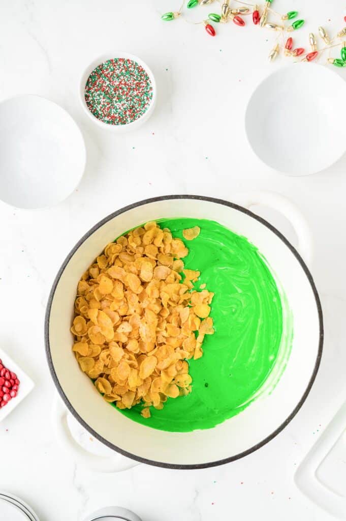 A pot with green melted marshmallow mixture with cornflakes being stirred in, and sprinkles in bowls nearby on the counter.