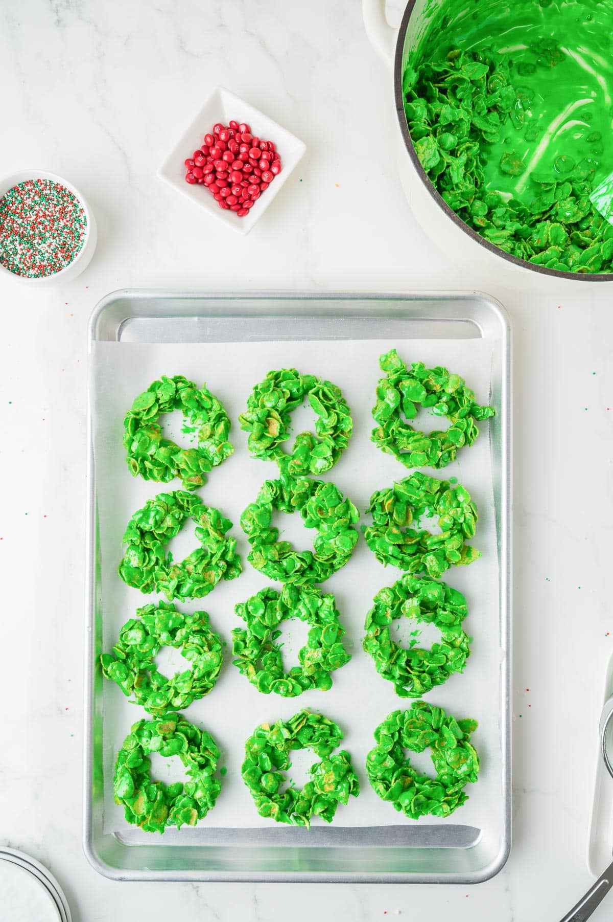 A baking sheet with twelve green cornflake wreath cookies,. A pot with green cornflake mixture and small bowls of sprinkles sit nearby on the counter.