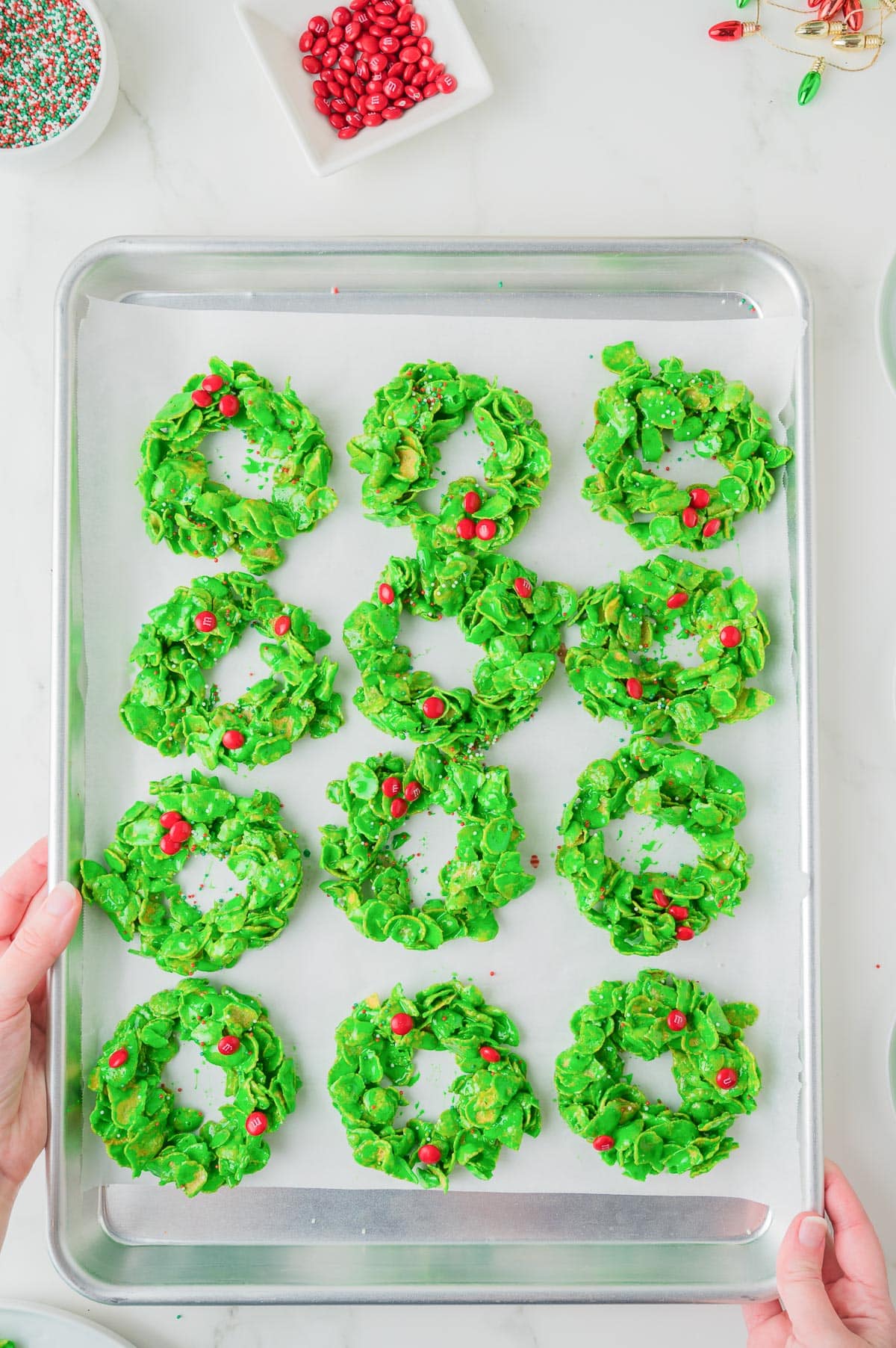 A baking sheet with twelve green cornflake wreath cookies topped with small red candies and sprinkles on parchment paper.