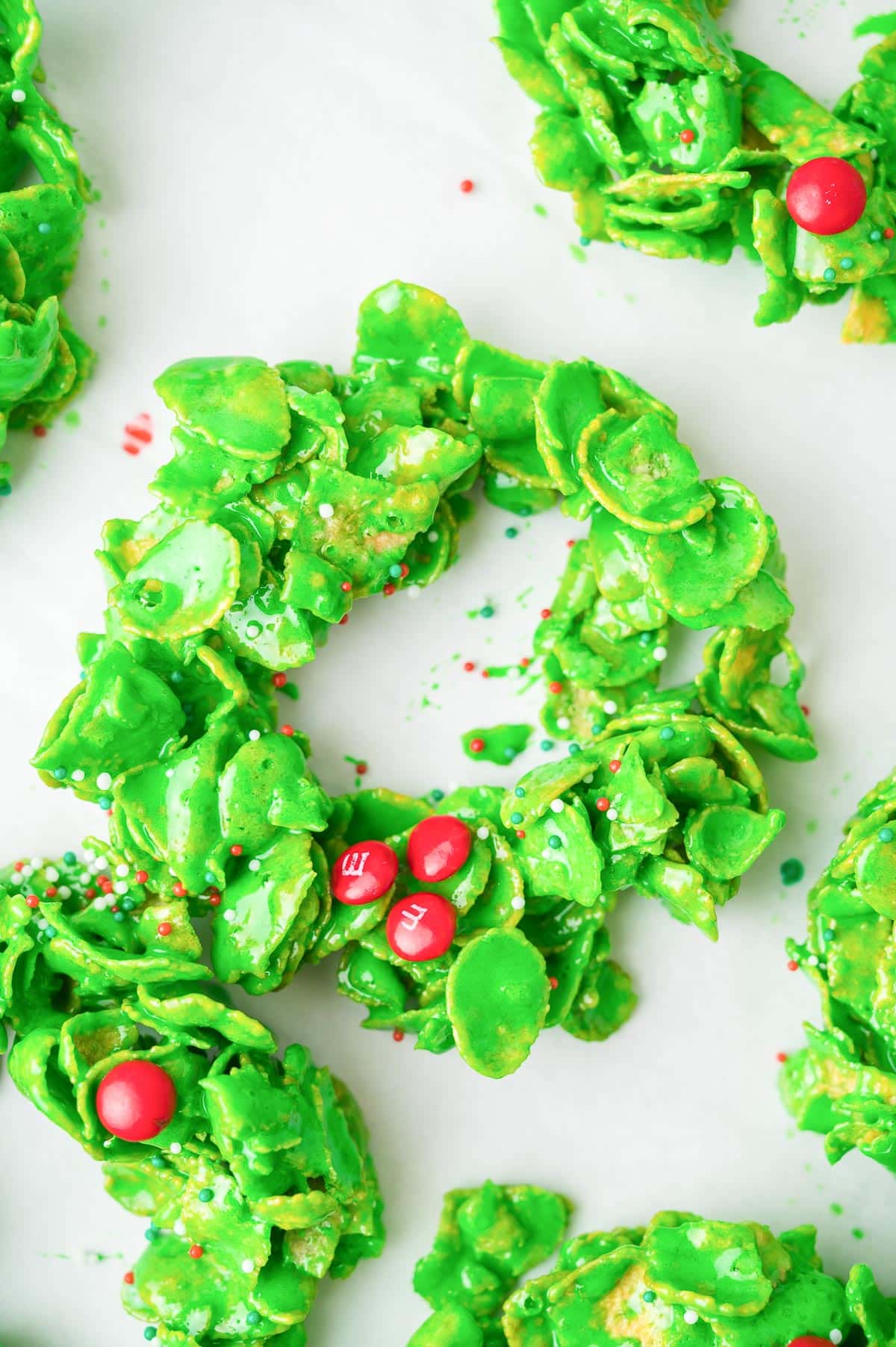 Green Christmas cornflake wreath cookies with red candy accents and sprinkles on a counter.