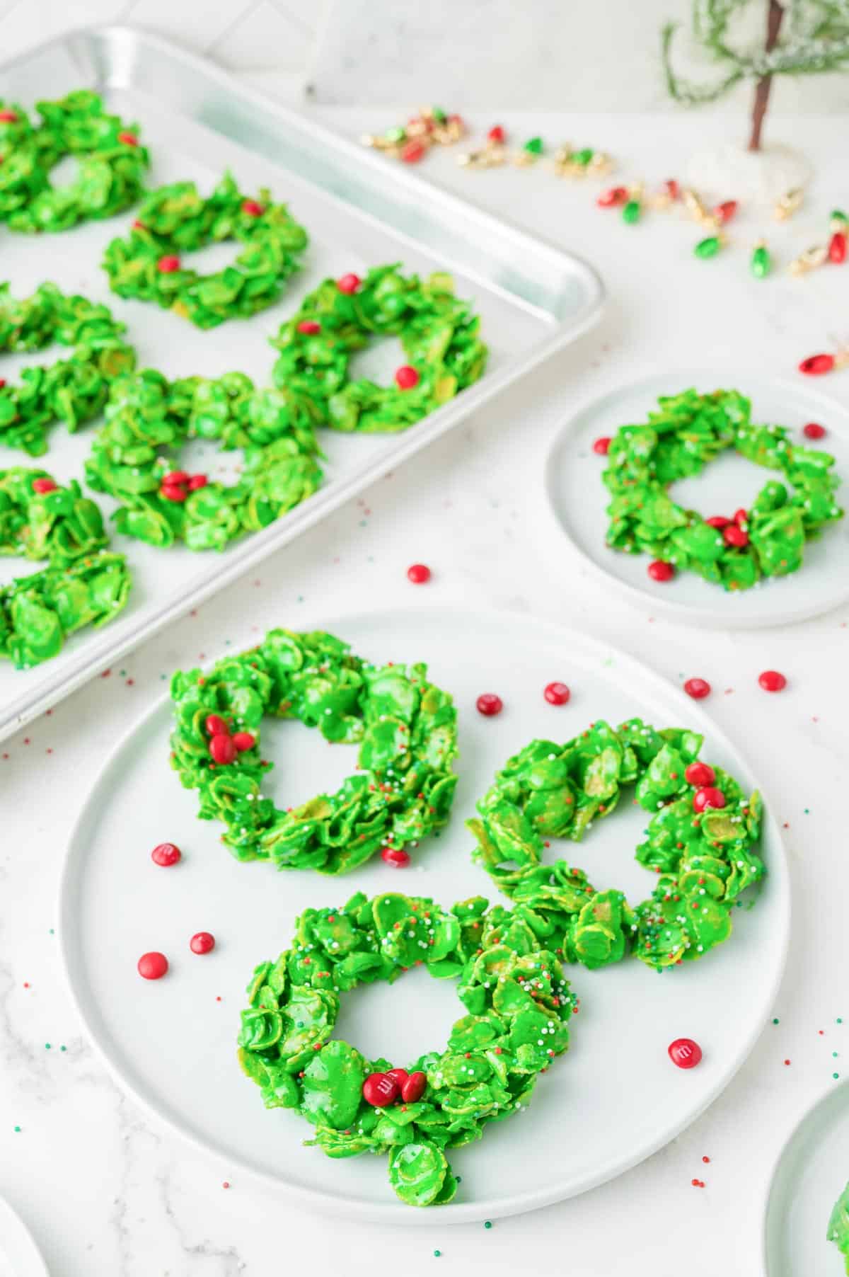 Green Christmas cornflake wreath-shaped cookies with red candies and sprinkles are arranged on white plates with a baking tray full in the background.