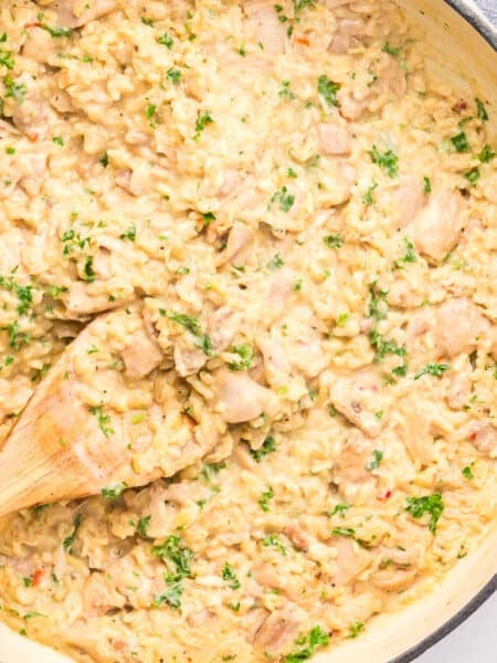 A pan of creamy chicken and rice, garnished with chopped parsley, with a wooden spoon resting on the side.
