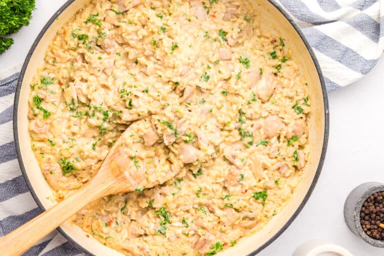 A pan of creamy chicken and rice, garnished with chopped parsley, with a wooden spoon resting on the side.