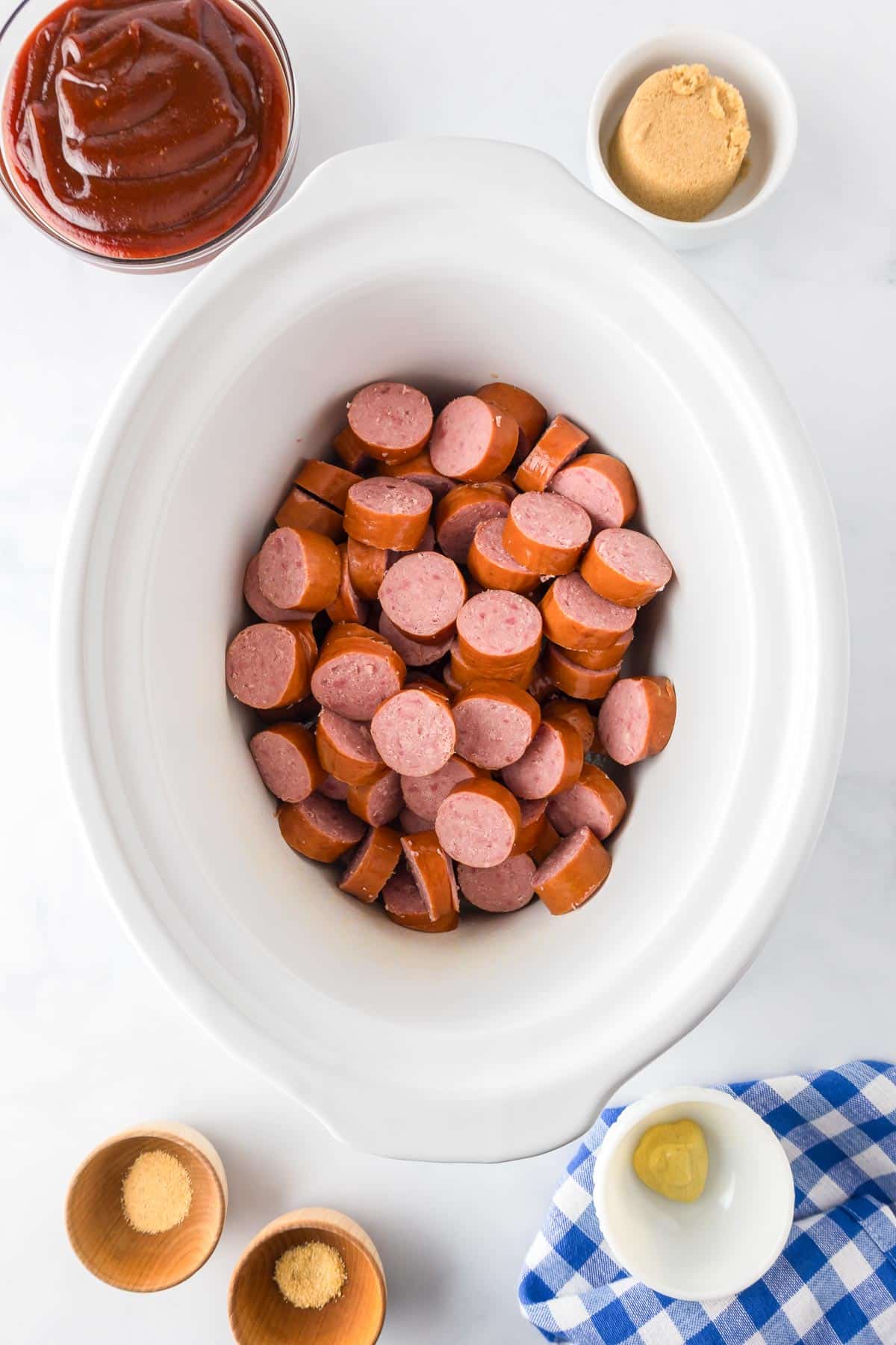 Sliced sausages in a white slow cooker surrounded by bowls of barbecue sauce, brown sugar, mustard, and seasonings.