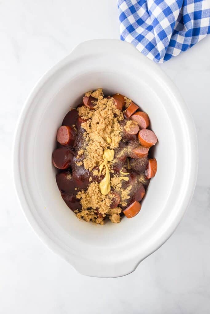 A white slow cooker filled with pieces of kielbasa, brown sugar, bbq sauce, spices and mustard on a counter.