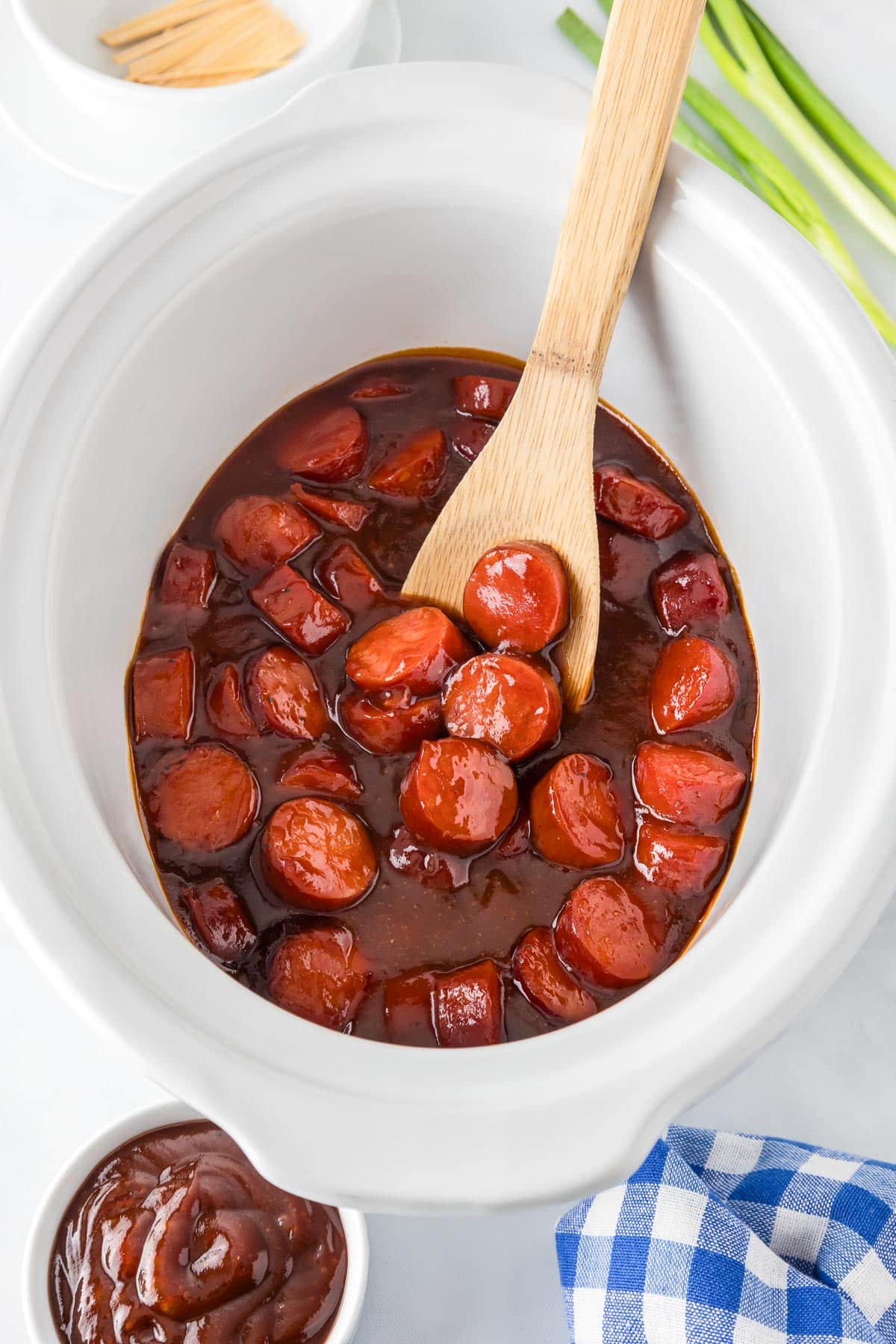 A slow cooker base filled with sliced kielbasa in a bbq sauce being stirred with a wooden spoon.