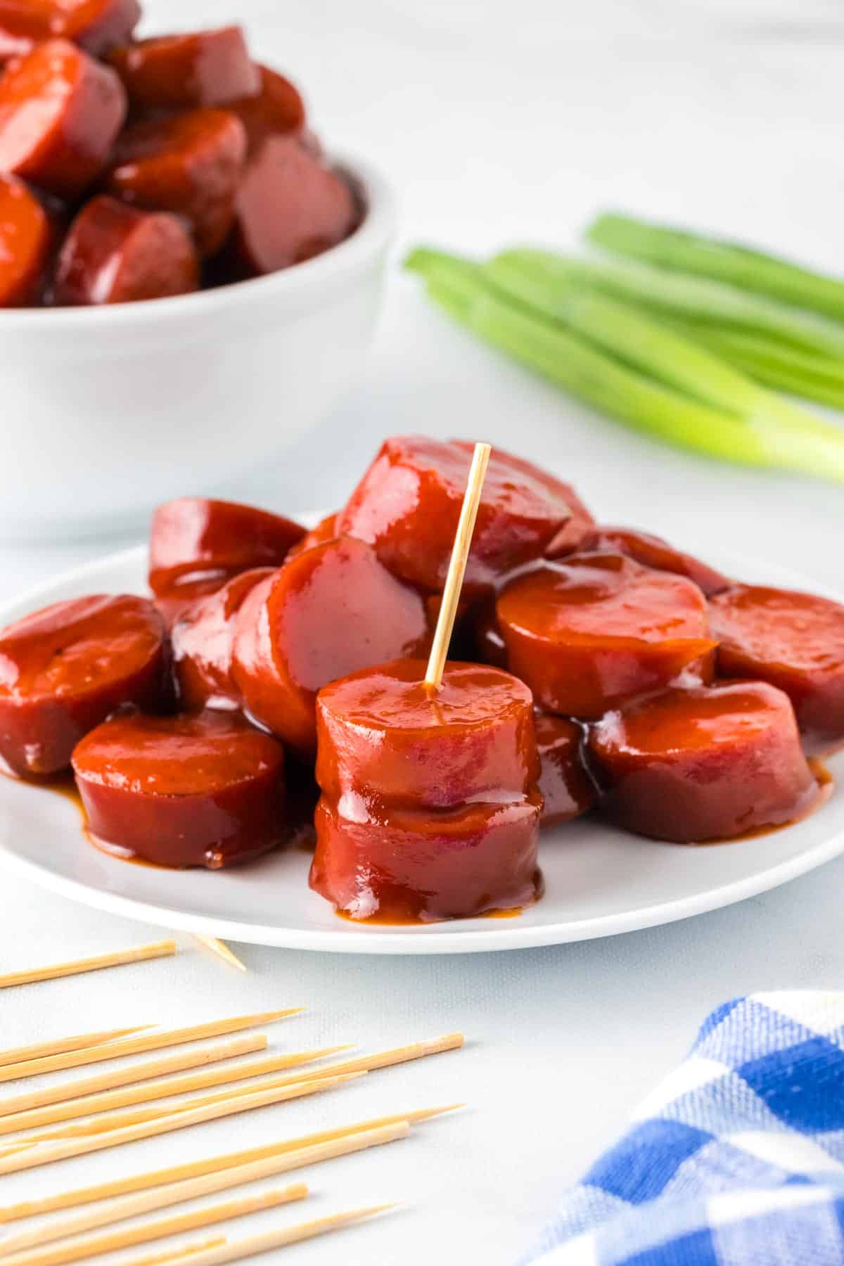 Plate of bbq kielbasa bites slices with a toothpick sticking out of two pieces in the front, and a second bowl of sausage bites and more toothpicks on the counter nearby.