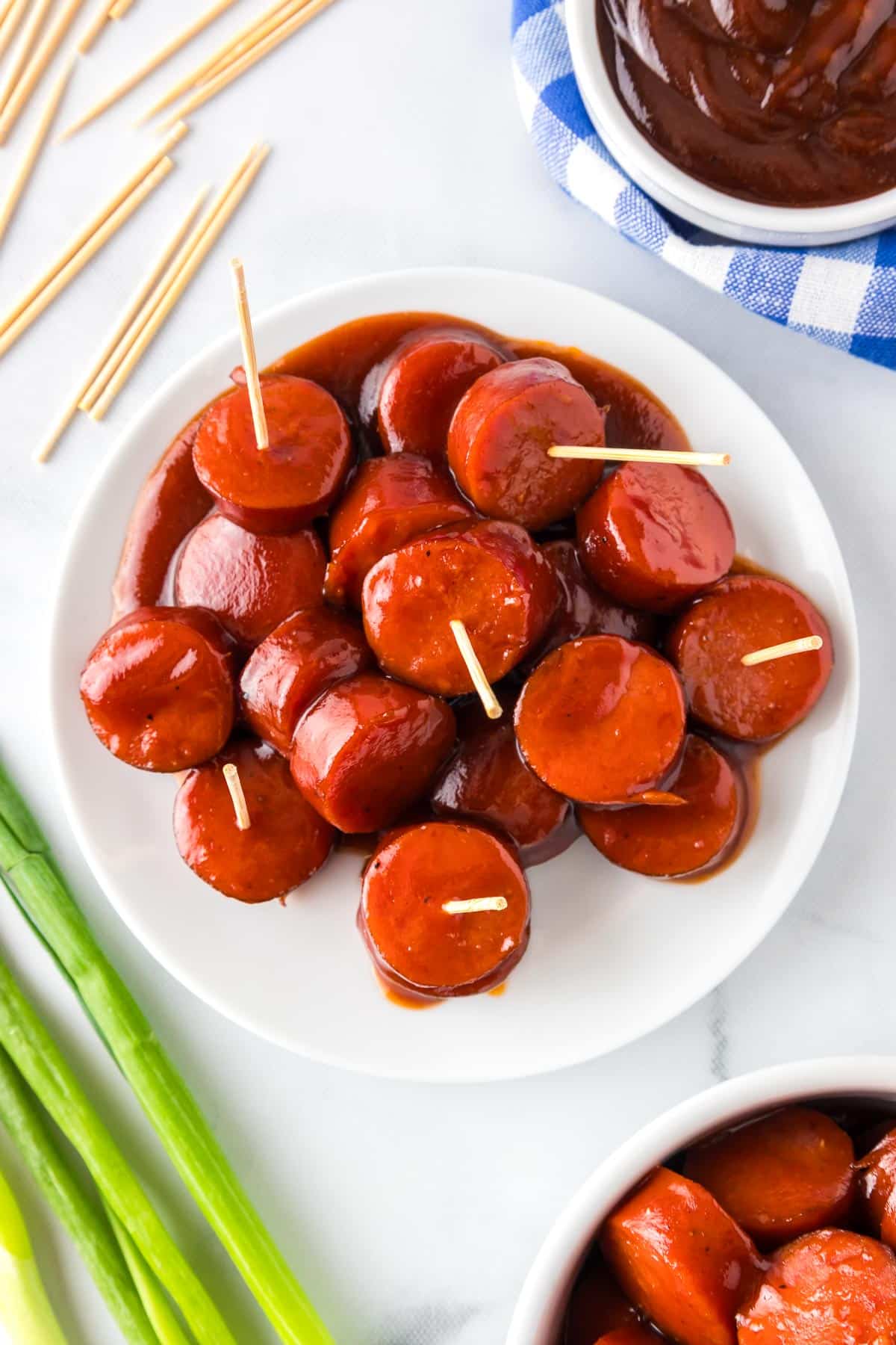 A plate of sliced sausages glazed in barbecue sauce with toothpicks sticking out of a few pieces from overhead with more toothpicks, another bowl of sausage and another bowl of bbq sauce nearby on the counter.