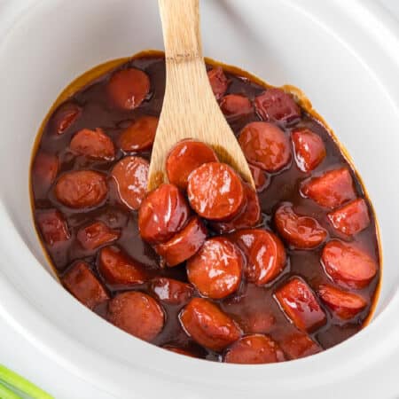 Sliced bbq smoked sausage bites in a barbecue sauce in a white crockpot being scooped with a wooden spoon.