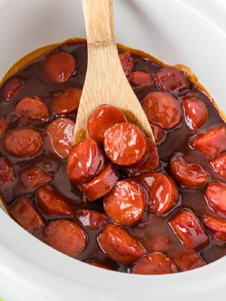 Sliced BBQ smoked sausage bites in a barbecue sauce in a white crockpot being scooped with a wooden spoon.