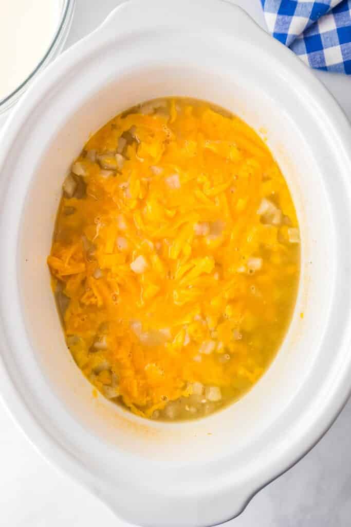 A white slow cooker filled with cooked potato hashbrowns, chicken broth and cheddar cheese with a bowl of milk nearby for crock pot hash brown potato soup.