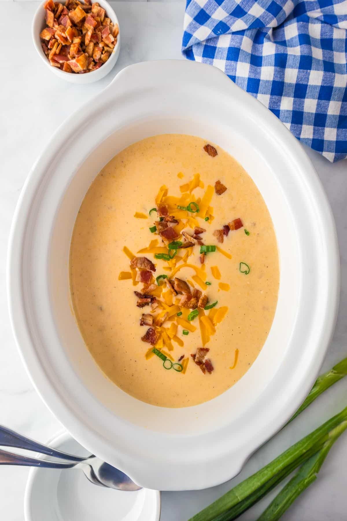 A finished crock pot full of creamy crock pot hash brown potato soup topped with bacon pieces, shredded cheese and green onions on a counter.