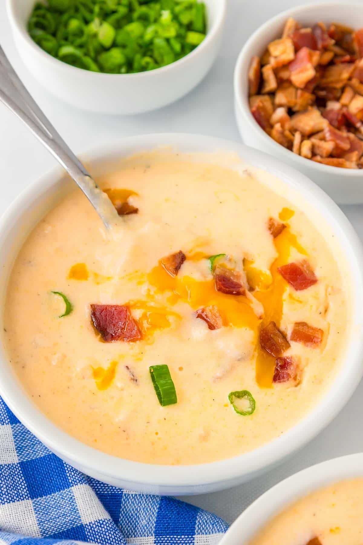 A bowl of creamy hashbrown potato soup topped with cheese, bacon pieces, and sliced green onions, with a spoon inside. scooping a bite.