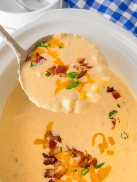 A ladle holding a scoop of creamy hash brown potato soup being scooped from a slow cooker base topped with bacon pieces, shredded cheese and green onion pieces.