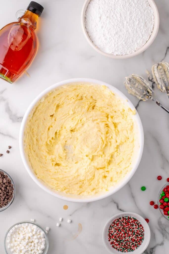 A bowl of butter and shortening beat together in a bowl with electric mixer beaters and other frosting for elf cupcakes in bowls nearby.