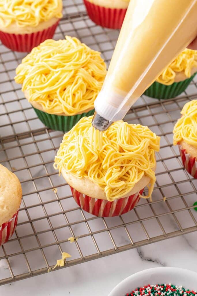 Piping yellow frosting onto cupcakes that resemble spaghetti for Buddy the Elf's favorite cupcakes on a cooling rack.