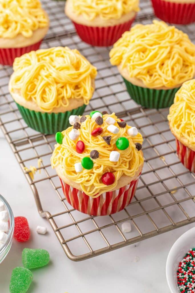 Several cupcakes with yellow frosting resembling spaghetti sit on a cooling rack. One cupcake is decorated with colorful candies and marshmallows like Buddy the Elf enjoys.