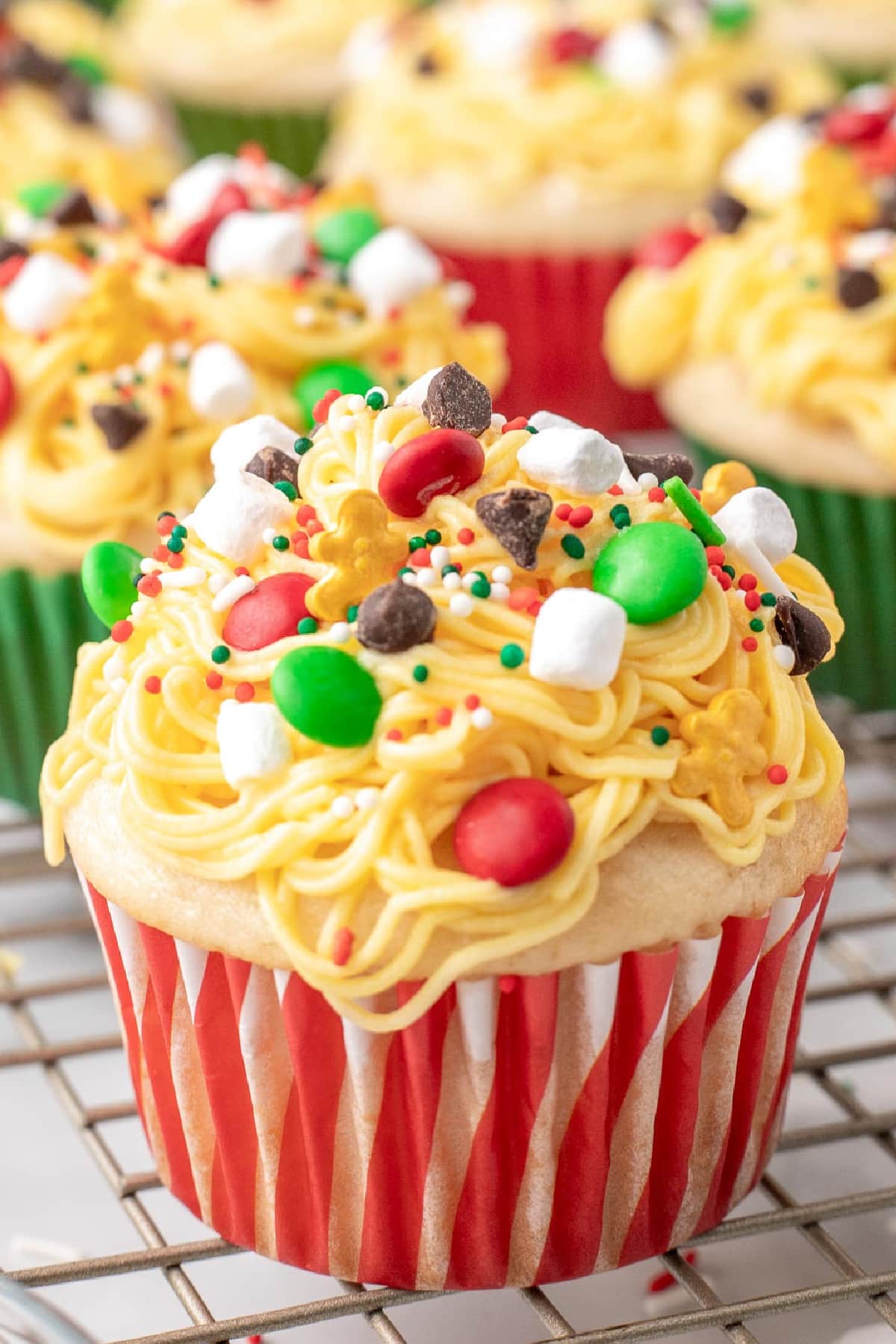 An Elf Christmas cupcake with more in the background, with yellow frosting made to look like spaghetti topped with candies, chocolate chips, holiday sprinkles and marshmallows.