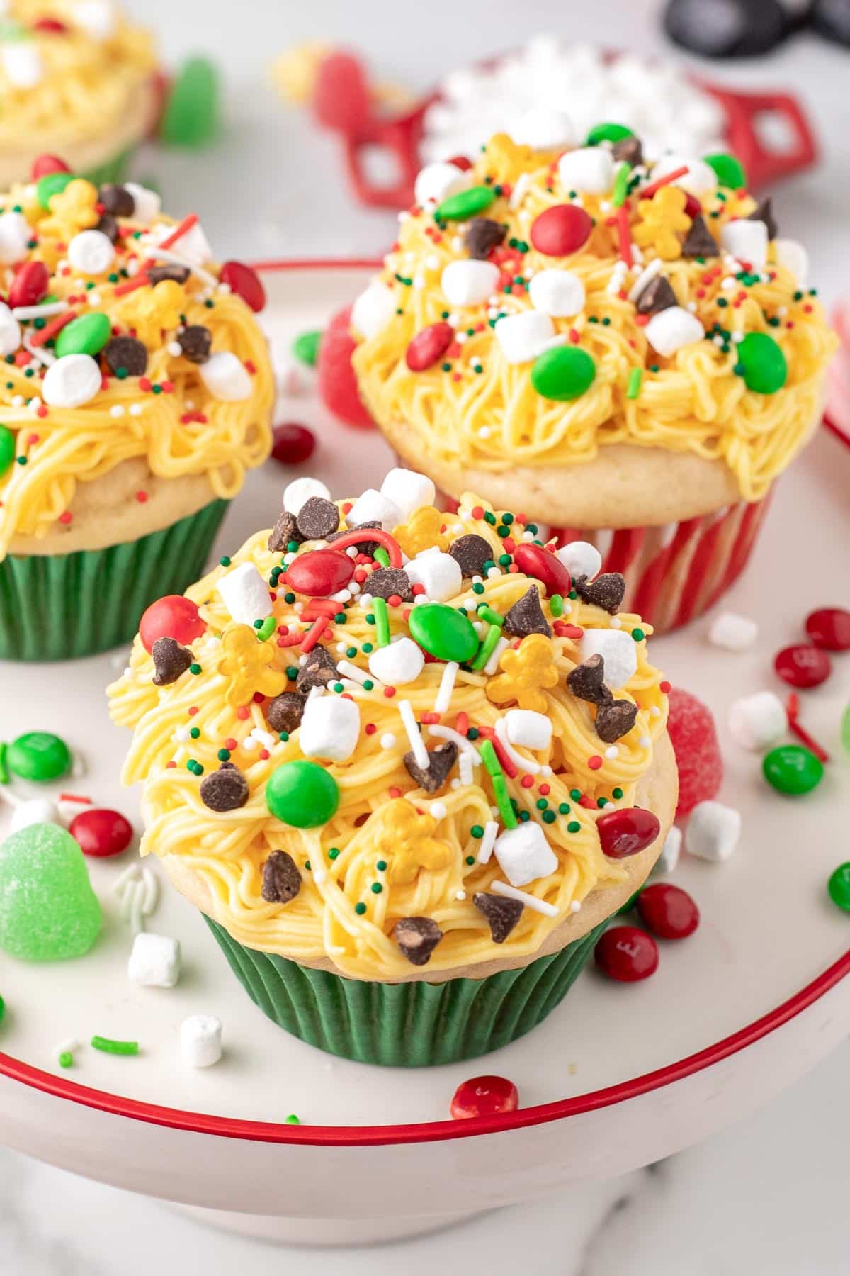 Elf Christmas cupcakes on a platter with yellow frosting made to look like spaghetti topped with holiday candies, sprinkles and chocolate chips.