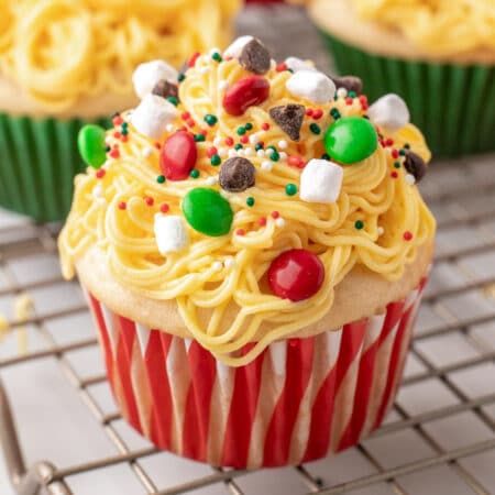 Close view of Elf Christmas cupcake on a wire rack with more in the background, with yellow frosting made to look like spaghetti topped with candies, chocolate chips, holiday sprinkles and marshmallows.