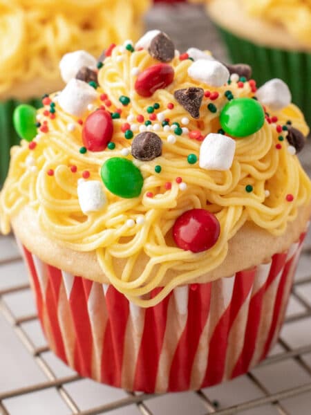 Close view of Elf Christmas cupcake on a wire rack with more in the background, with yellow frosting made to look like spaghetti topped with candies, chocolate chips, holiday sprinkles and marshmallows.