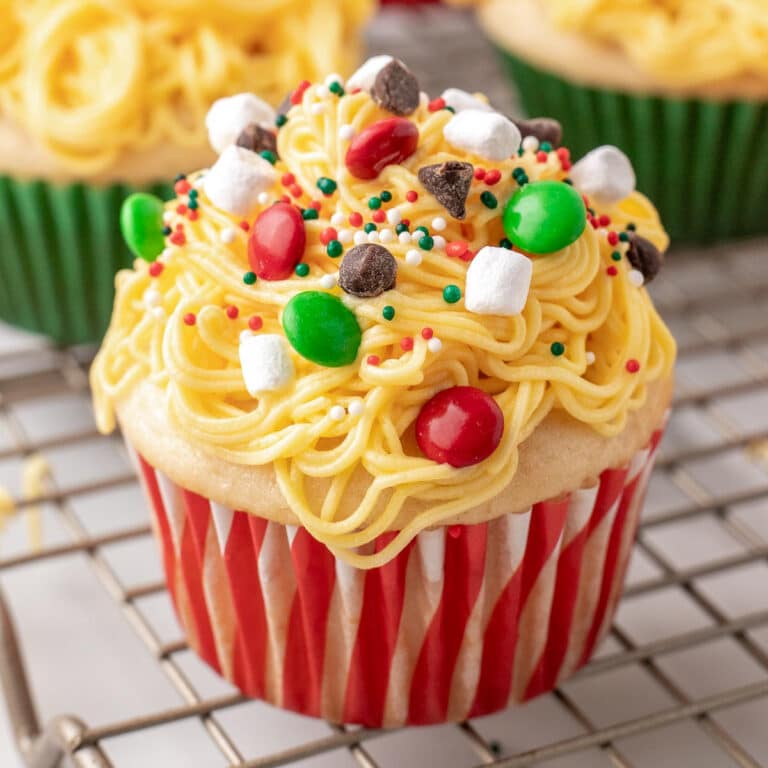 Close view of Elf Christmas cupcake on a wire rack with more in the background, with yellow frosting made to look like spaghetti topped with candies, chocolate chips, holiday sprinkles and marshmallows.