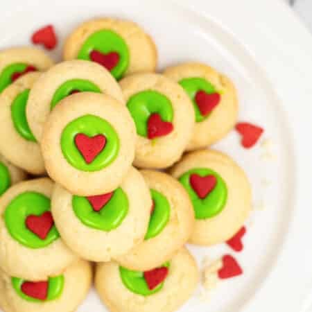 A plate of Grinch thumbprint cookies with green icing and red heart decorations on a plate from overhead.