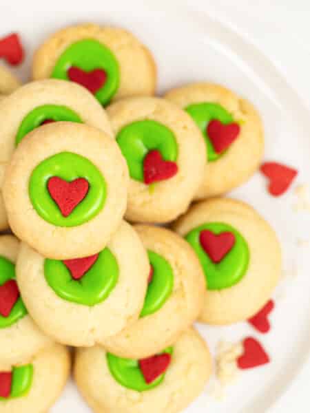 A plate of Grinch thumbprint cookies with green icing and red heart decorations on a plate from overhead.