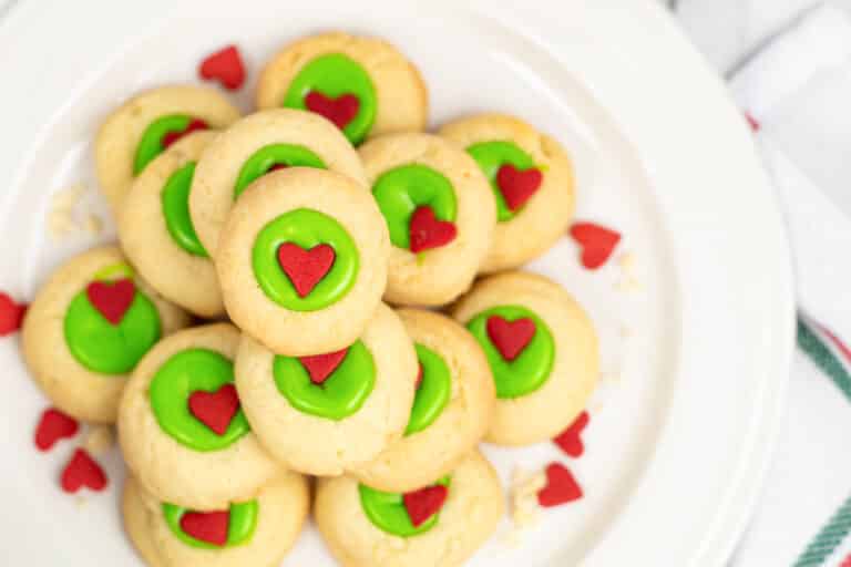 A plate of Grinch thumbprint cookies with green icing and red heart decorations on a plate from overhead.