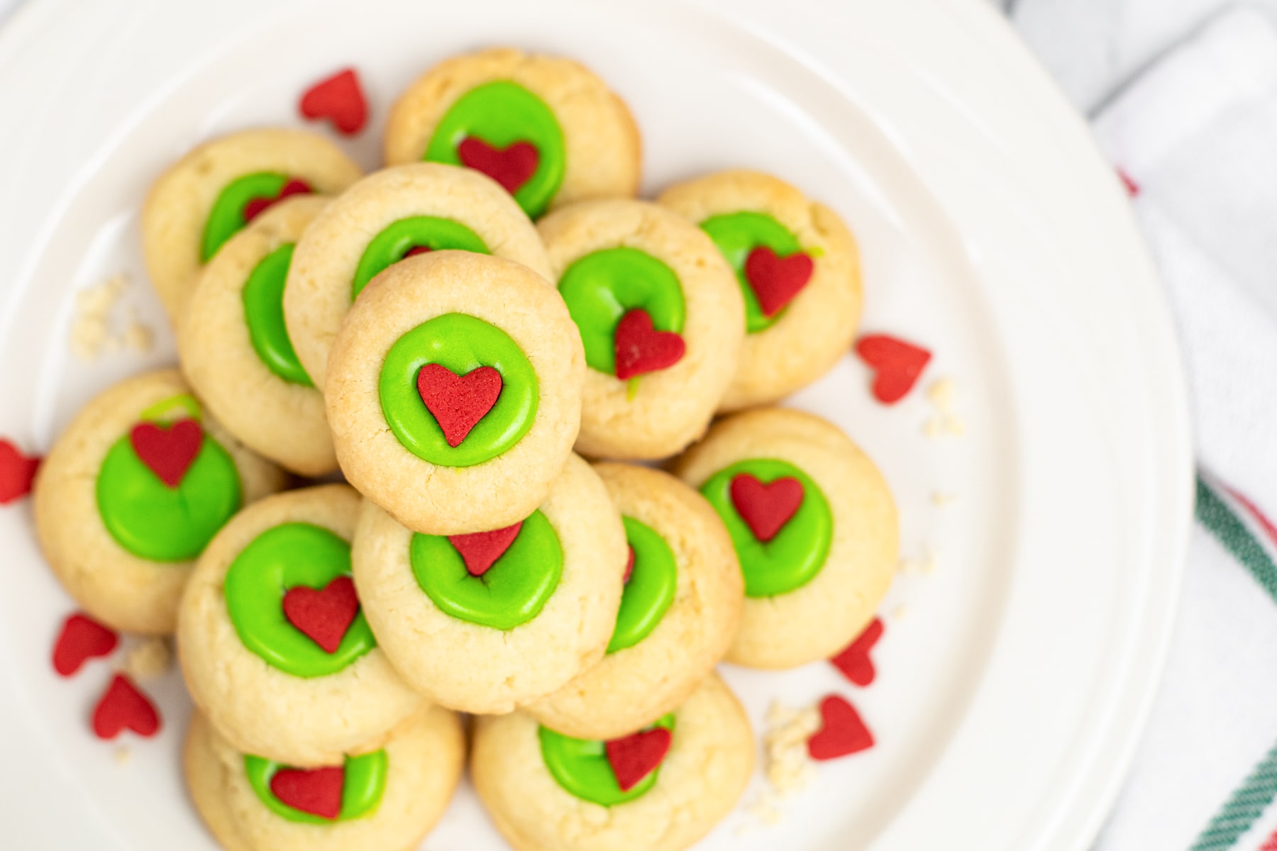 A plate of Grinch thumbprint cookies with green icing and red heart decorations on a plate from overhead.