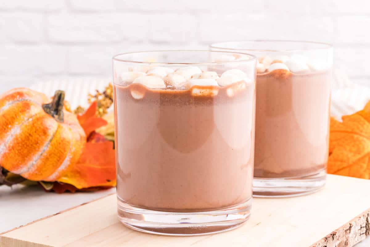 Two glasses of mexican hot chocolate topped with marshmallows on a wooden surface, with small pumpkins and autumn leaves in the background.