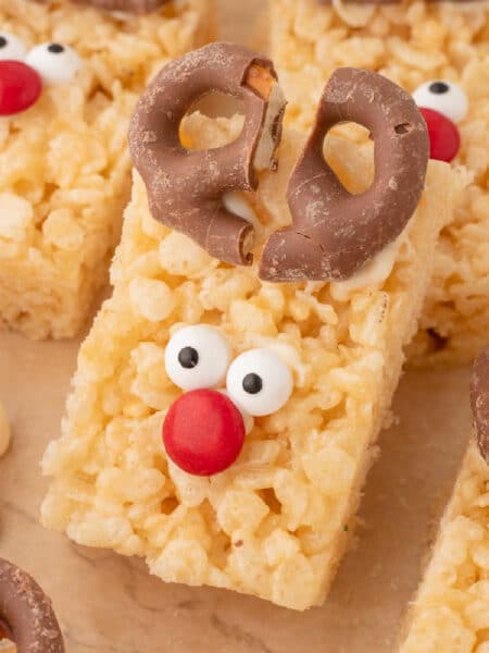 Close up of several stacked rectangular Rice Krispie treats decorated as reindeers with chocolate-covered pretzel antlers, candy eyes, and red candy noses.