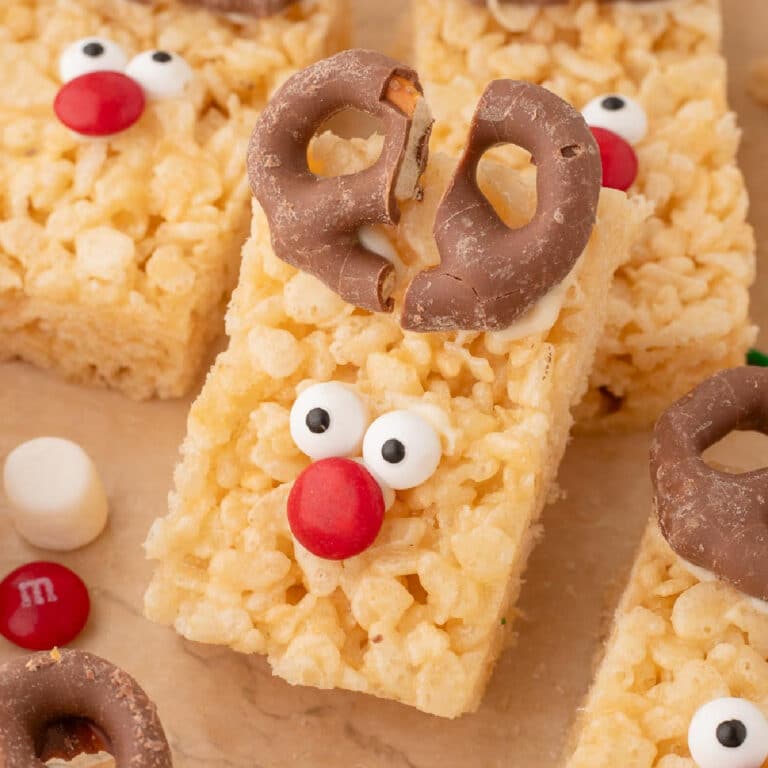 Close up of several stacked rectangular Rice Krispie treats decorated as reindeers with chocolate-covered pretzel antlers, candy eyes, and red candy noses.