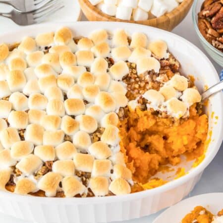 Sweet potato casserole topped with toasted marshmallows in a white dish, partially served, with bowls of pecans and marshmallows nearby.
