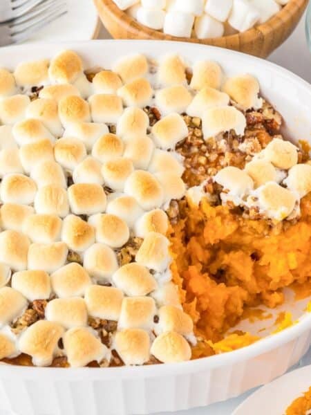 Sweet potato casserole topped with toasted marshmallows in a white dish, partially served, with bowls of pecans and marshmallows nearby.
