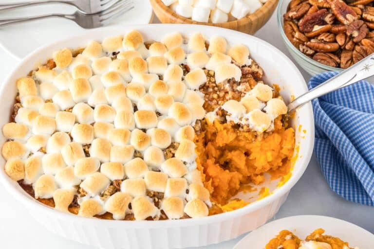 Sweet potato casserole topped with toasted marshmallows in a white dish, partially served, with bowls of pecans and marshmallows nearby.