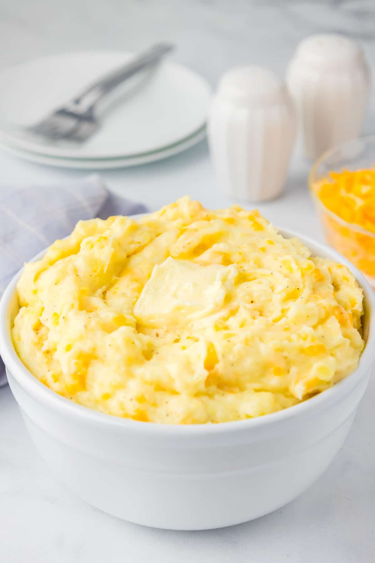 A large bowl of yellow Yukon Gold mashed potatoes with a pat of melty butter on top, with plates, forks, a bowl of shredded cheese and salt and pepper shakers in the background.