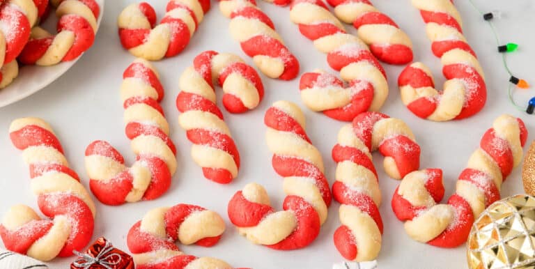 Wide view of red and white twisted candy canes cookies shaped like candy canes and sprinkled with sugar spread out on a counter.