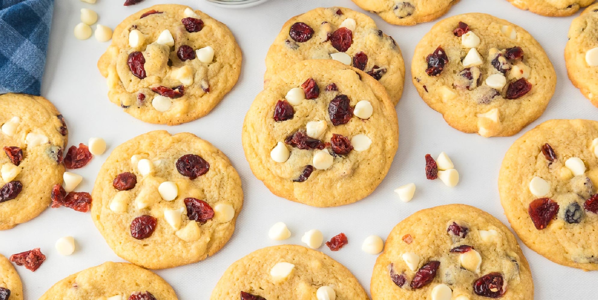 A batch of white chocolate chip cranberry cookies spread out over a counter with a few loose cranberries and white chocolate chips.