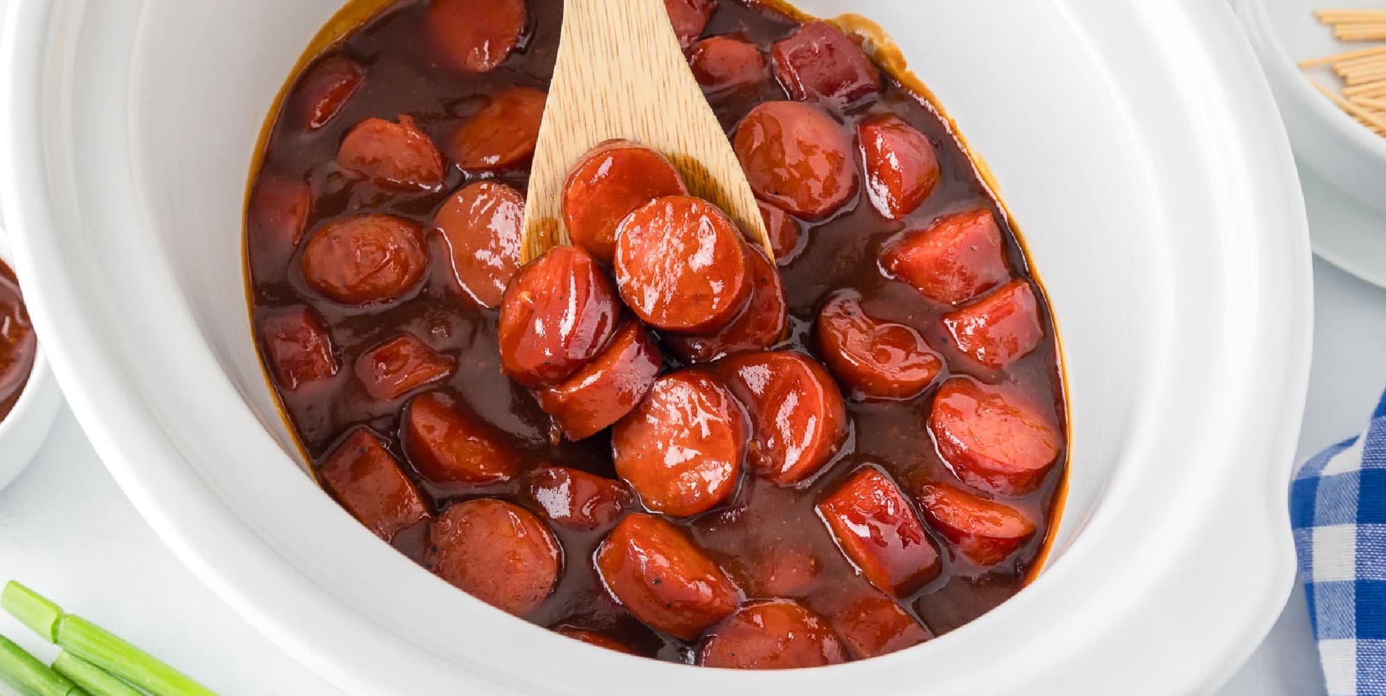 A wide view of a wooden spook scooping sliced bbq smoked sausage bites in a barbecue sauce in a white crockpot.