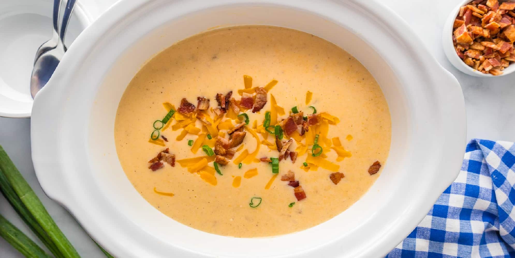 Wide view of crock pot hashbrown potato soup in a slow cooker base topped with bacon, cheddar cheese and green onions on a table.