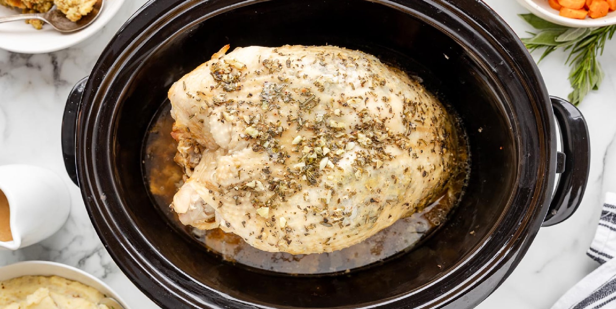 Wide view of a seasoned bone in turkey breast cooking in a slow cooker surrounded by various side dishes in bowls on a counter.